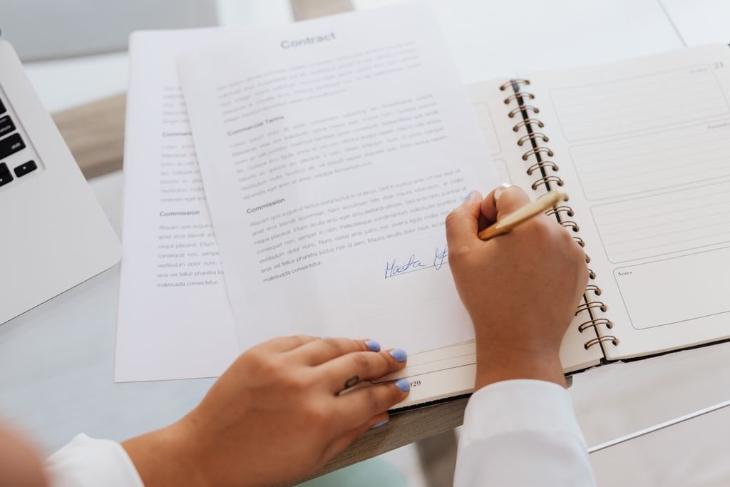 A hand signing a printed contract beside an open notebook