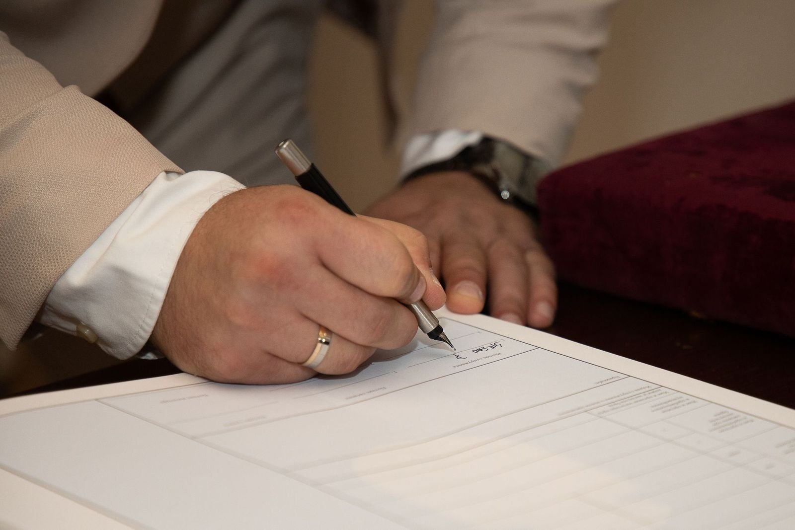 A hand with a white shirt cuff and a ring signing a document with a fountain pen