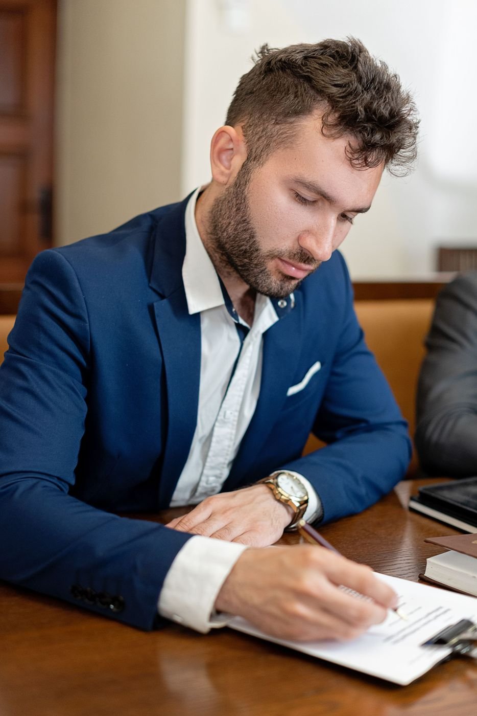 A man in a blue suit signing a document at a wooden desk