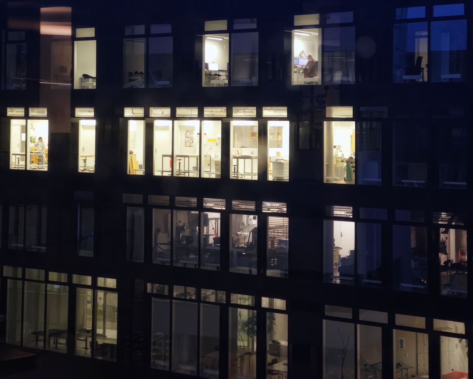An office building at night, rows of lit windows with desks and lamps inside