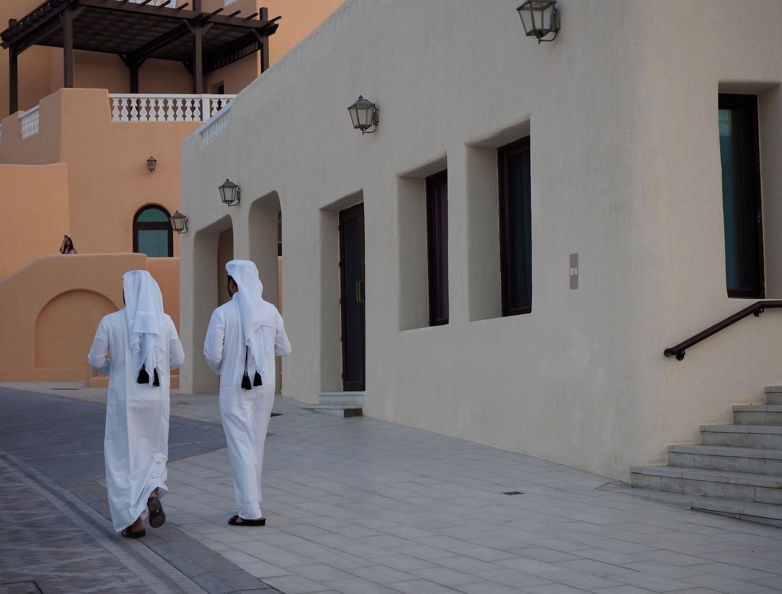 Two men in white kandura walking along a quiet street past a pale building